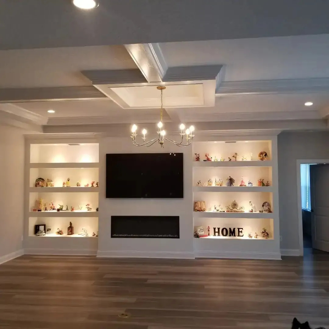 White built-in shelving with lighted niches around a TV and electric fireplace under a coffered ceiling