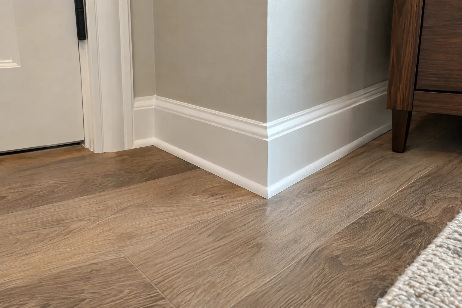 Thin white base shoe along the bottom of a painted baseboard where it meets light oak flooring near a door casing